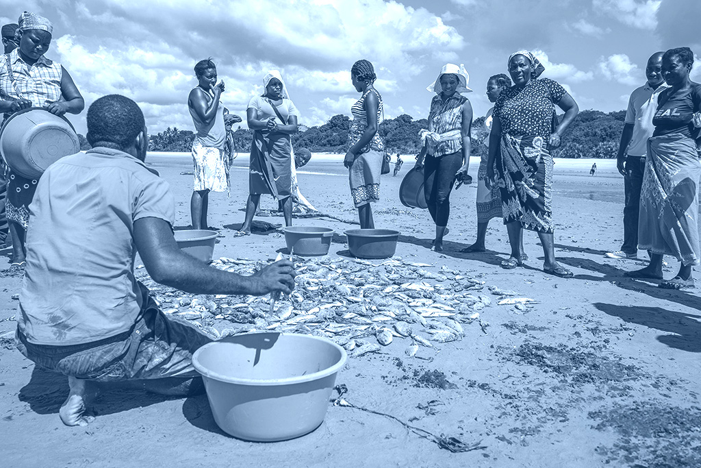 Women from a fishing community reviewing catch data sheets on a beach beside fishing boats